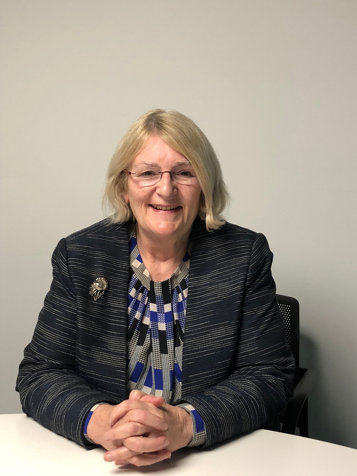 Photograph of Linda Gilroy sat down behind a table with hands clasped in front of her. She is wearing a blue, black, grey and white patterned top and a stripy patterned jacket with a brooch.
