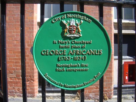 Green plaque from the City of Nottingham on black bars outside St Mary's Churchyard. Plaque reads St Mary's Churchyard, burial place of George Africanus (1763-1834), Nottingham's first Black entrepreneur.
