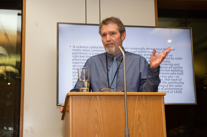 Professor Chris Given-Wilson stands at a wooden lectern in front of a microphone. He is mid speech with his left hand gesturing and is standing in front of a screen with an illegible quote written on it. He is wearing a grey shirt with grey tie.