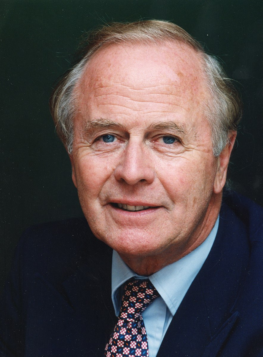 Photograph of bust and head of Sir John Hannam. He is looking forward with thinning, greying hair. He has blue eyes and a slight smile. He is wearing a blue shirt, dark suit jacket, and a checked tie.
