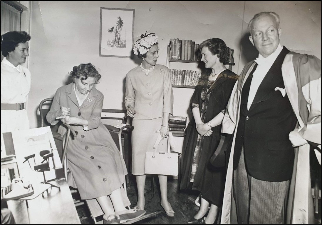 A black and white photograph of five people. On the far left is a female nurse with dark hair pinned up and wearing a white uniform with belt, next is a woman sat in a wheelchair with dark curled hair and wearing a long buttoned coat and shoes, next is a woman stood pointing, she is wearing a hat, skirt suit, necklace and holding a bag, next a woman with short dark hair, wearing academic robes and a dress, on the end right is a man with thinning, light hair, wearing a suit with academic robes. They are in a room with book shelves and a painting.