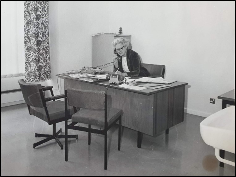 A black and white photograph of Margaret Agerholm sat behind a desk answering the phone. She has light, short hair and is wearing a light coloured top and dark jacket. The room has a desk, two chairs, and a cabinet. The desk is filled with lots of papers.