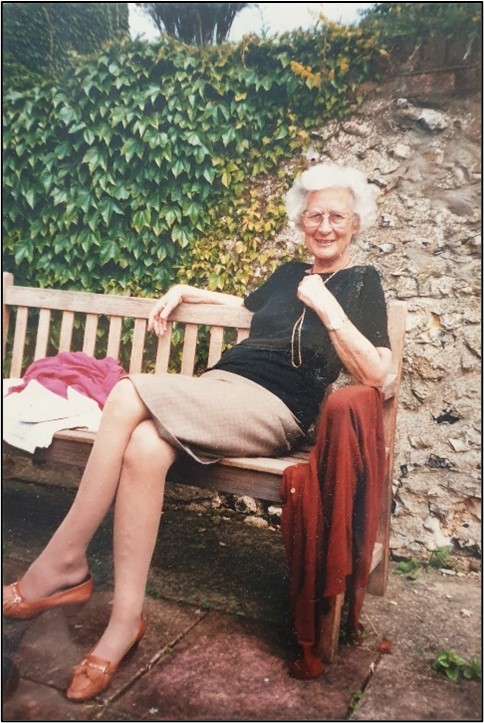 A full body photograph of Dr Margaret Agerholm sat down on a bench in front of a stone wall that is wrapped in ivy. Agerholm is smiling, her left hand is touching her necklace and her right arm is resting on the back of the bench. Her legs are crossed. She is bearing a black top, beige skirt, and brown shoes.