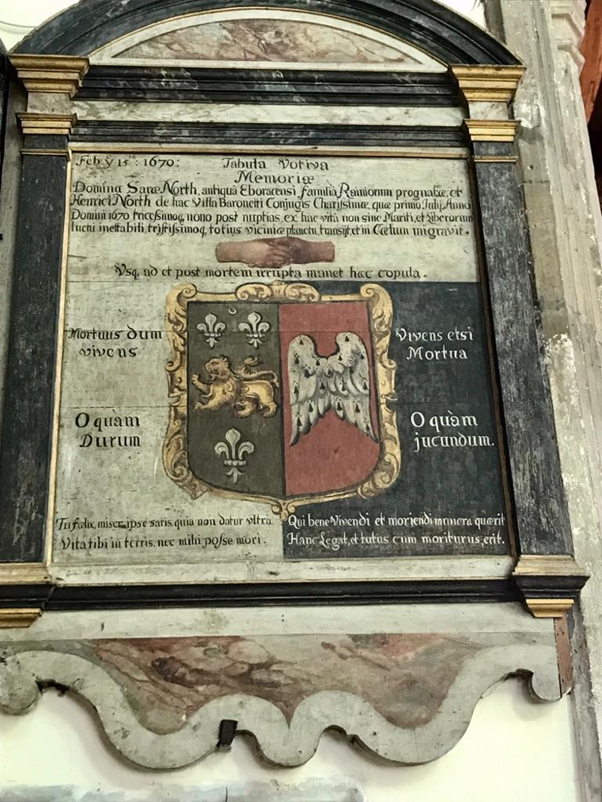 A monument on a chapel wall. It is framed by a black marble effect with gold accents. Inside there is family arms - the coat is split in two, on the left it is green with a golden lion and on the right it it red with white wings, it is framed in gold. Above the coat of arms are two hands clasped. Around this is writing in Latin.