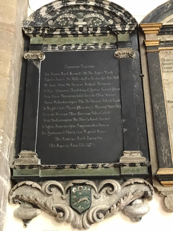 A memorial on a chapel wall. It is a black wooden tablet. At the top is a black and white marble design. On it is writing in Latin. Due to picture quality it is not readable. At the bottom is a coat of arms. It is green and has a lion on it.