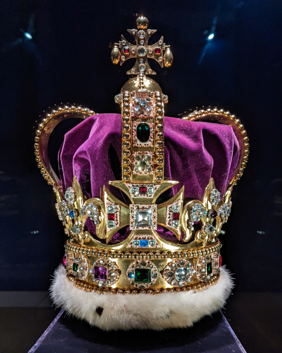 A photograph of a crown. The crown is composed of a solid gold frame, set with tourmalines, white and yellow topazes, rubies, amethysts, sapphires, garnet, peridot, zircons, spinel, and aquamarines, step-cut and rose-cut and mounted in enamelled gold collets, and with a velvet cap with an ermine band. The band of the crown is bordered by rows of gold beads and mounted with sixteen clusters, each set with a rectangular or octagonal step-cut stone in a collet decorated in enamel with modelled acanthus leaves, surrounded by rose-cut topazes and aquamarines, mainly round. Above the band are four crosses-pattée and four fleurs-de-lis mounted with clusters of large step-cut stones and smaller rose-cut stones. The two arches are mounted with gold beads and applied mounts with enamelled settings, containing step-cut stones and clusters of rose-cut smaller stones.