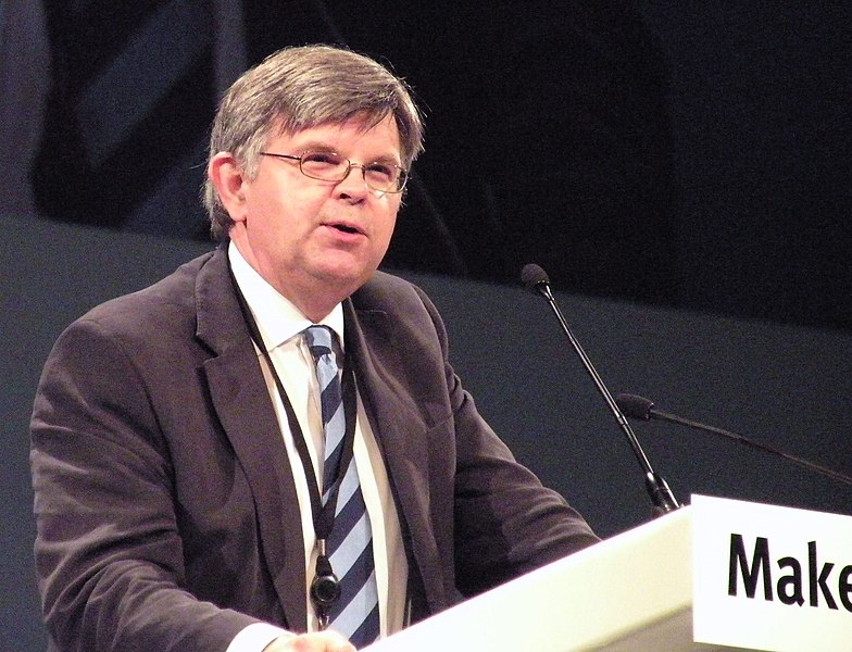A photograph of a white man with greying hair stood behind a lectern that has two microphones on it. He has glasses and is wearing a white shirt, blue striped tie, a grey jacket, and has a lanyard on. The background is grey.