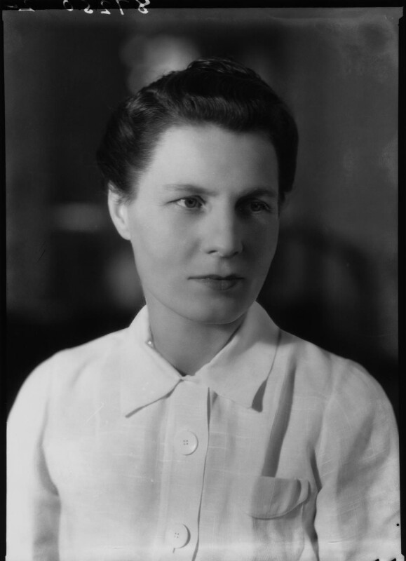 Black and white photograph of a white woman looking left of centre. She has dark, pinned back hair, and is wearing a white shirt. The top half of her body and head are in the photograph. The background is blurred and dark.