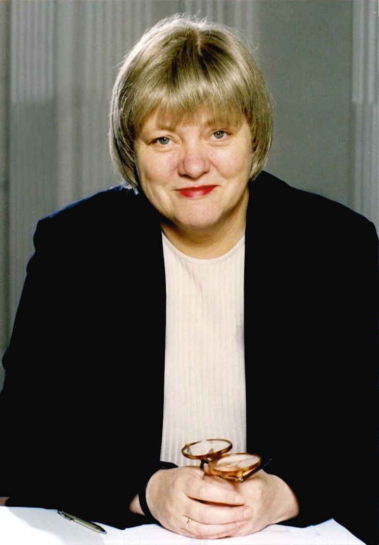 A photograph of a white woman with a short dark blonde bob. Sh is wearing red lipstick, a dark blazer and a light top. She is holding glasses in both her hadns and is sat behind a white table.