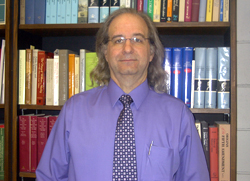 A photograph of a white man with shoulder length light hair wearing glasses. He is wearing a blue/purple shirt and a patterned tie with the colours of white and purple. There is a pen in his shirt pocket and a bookcase in the background.