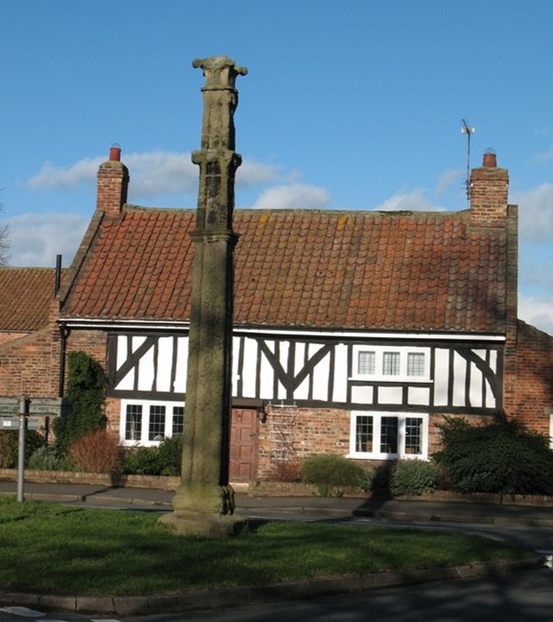 A photograph of a memorial. It is in the shape of a pillar. The memorial is on a patch of grass next to a road. Behind the memorial is a two-storey house, part of which is in a mock-Tudor design. The sky is blue with a few clouds passing behind the house.