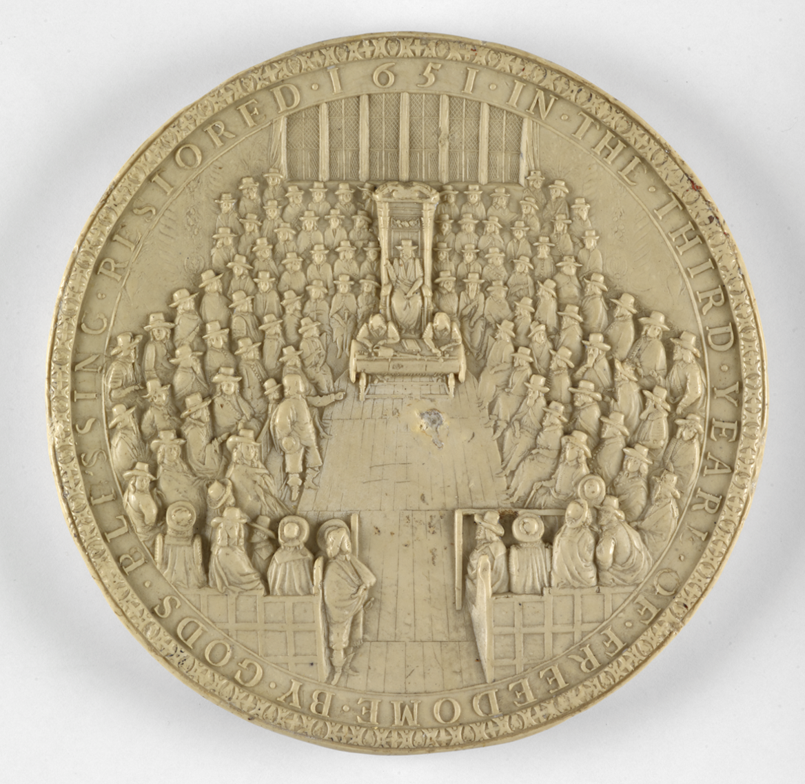 A cast of a seal. It is circular. There are people sat in the centre looking inwards - it is the House of commons. The Speaker is sat on a large chair in the centre. There is writing around the outside. 