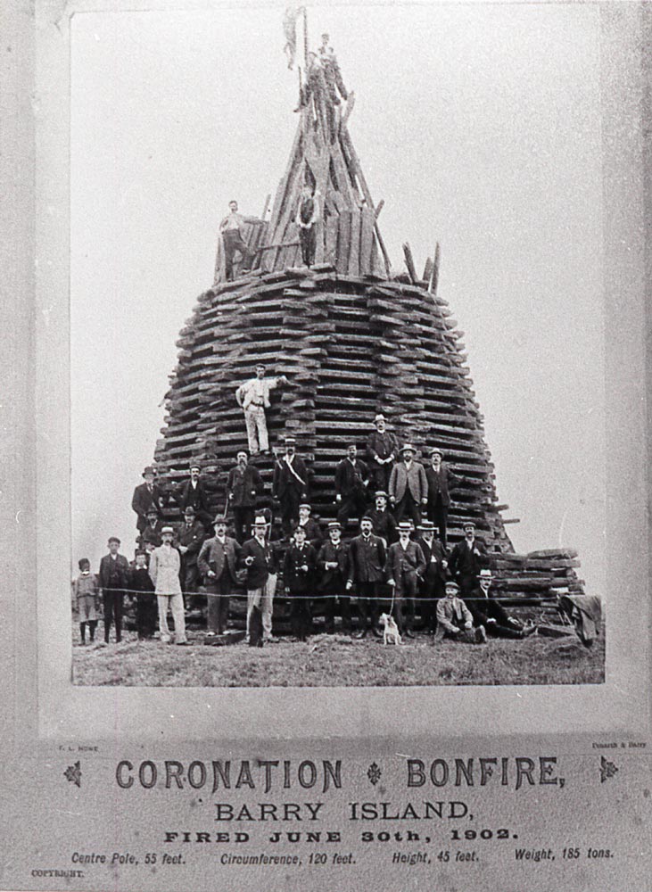 And image of a very high bonfire (unlit) there are people sat on top of it and more people stood around the bottom of it. There is writing at the bottom of the image: Coronation bonfire, Barry Island, fired June 30th, 1902. Centre pole: 55 feet. Circumference: 120 feet. Height: 45 feet. Weight: 185 tons.