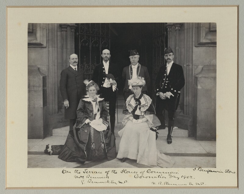 A black and white photograph of six white people. Two women are sat on chairs in front wearing formal dresses and hats. Four men are stood behind them wearing shirt, jacket, and trousers/ Two are wearing a hat, one is holding his hat and gloves in his hand.