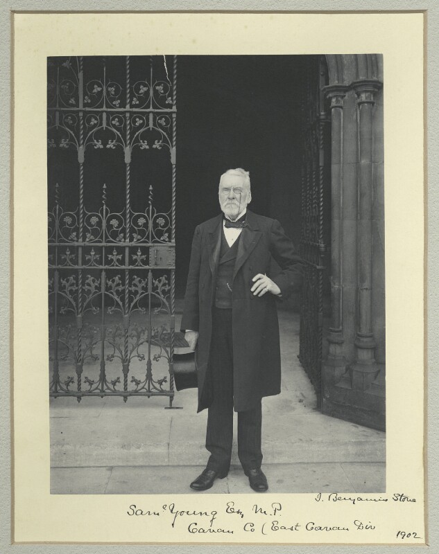 A photo of an older white man stoof in front of a doorway. He is wearing a white shirt, dark waistcoat, dark jacket dark trousers, and a bowtie. He also has a pocket watch, a monocle and is holding a top hat in his right hand.