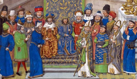 A painting of the coronation of Richard II who is in the centre of the image in blue wearing a crown. There are men in different attires to his left and right. In front of him is a bishop wearing white, blue, green and gold patterned robes and a white and gold hat.