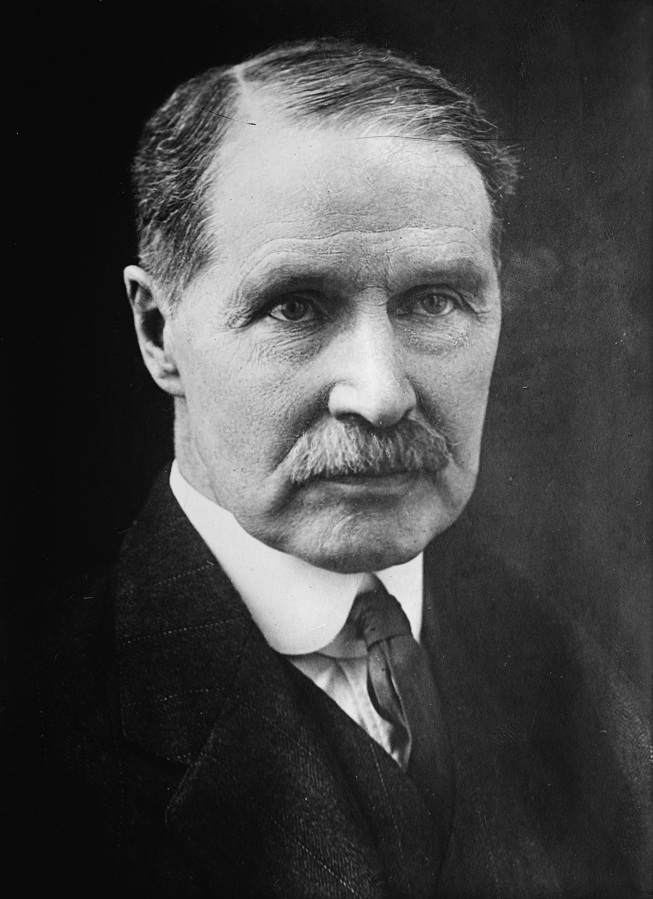 A photograph of a white man. His shoulders and head are in frame. He has thinning, dark hair and a dark moustache. He is wearing a 3 piece dark suit, white shirt, and dark tie.