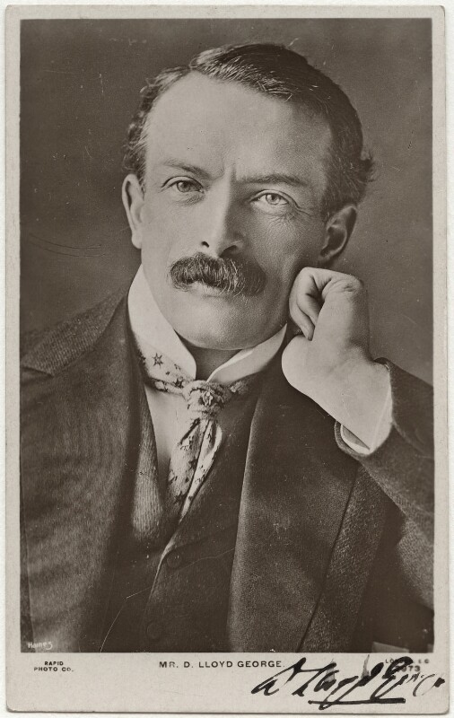 A black and white photograph on a postcard of the shoulders and head of a white man. He is wearing a 3 piece suit and is leaning on his left hand. He has dark short hair and a moustache.