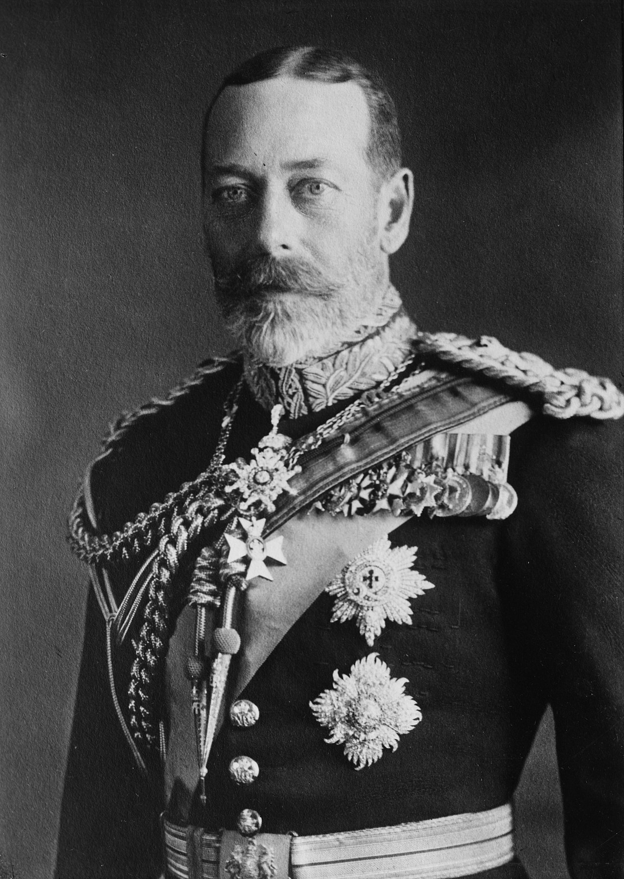 A black and white photograph of a white man, George V, stood. His torso and head are in frame. He is wearing military uniform and has medals pinned to his chest. He has short dark hair and a greying beard.