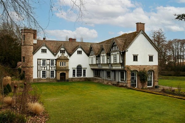A photograph of a L shaped half timbered building with three gables. it is surrounded by a mowed lawn and some trees.