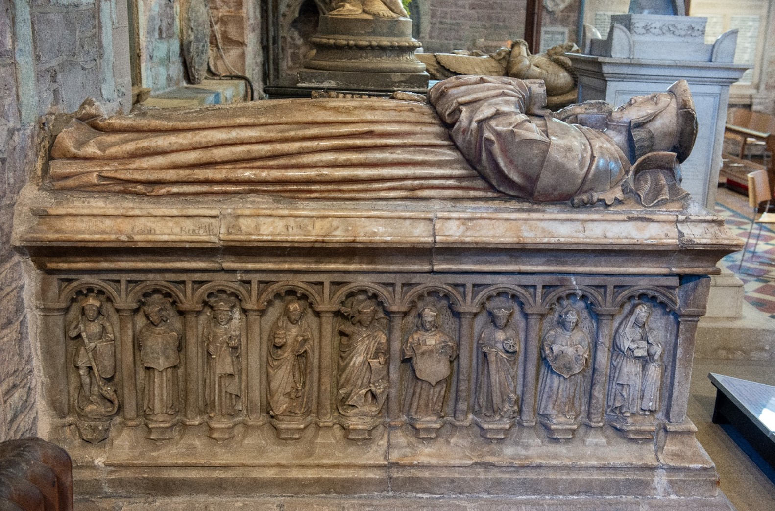 A tomb with two figures lying on top. Below these figures on a side panel are 9 figures.
