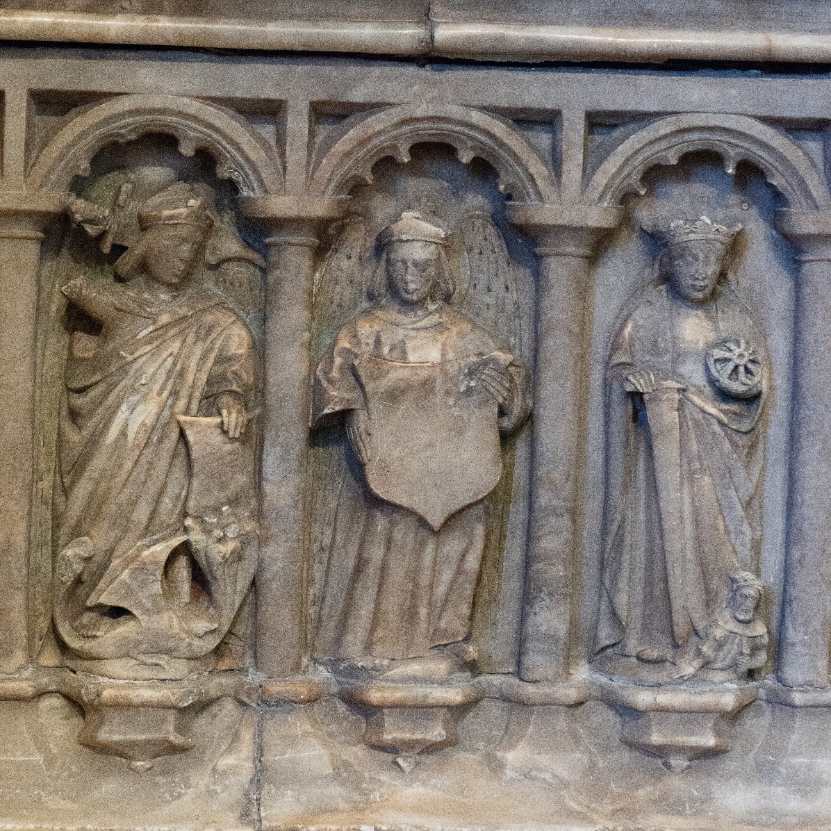 A panel of a tomb showing three figures. One is slaying a dragon, one is holding a shield and one is holding a wheel.