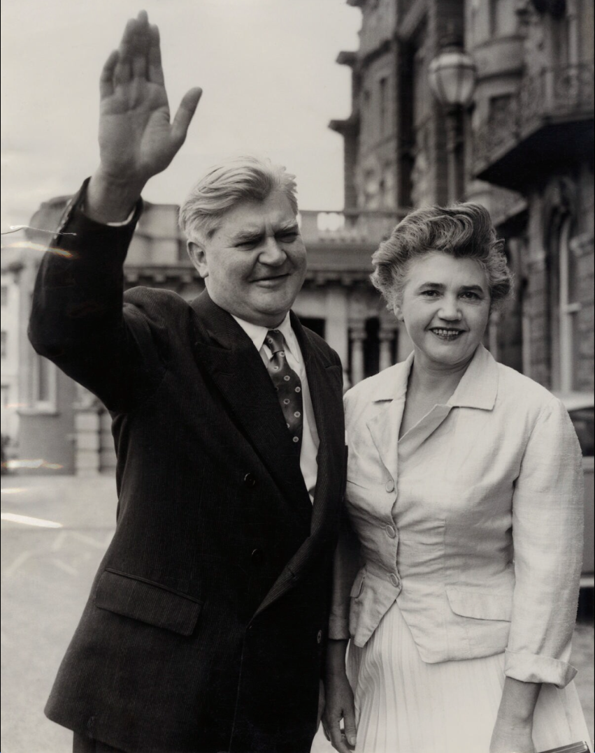 A photograph of a white man and white woman stood next to each other outside. They are close to each other. The man is waving his right hand. They are both smiling.
