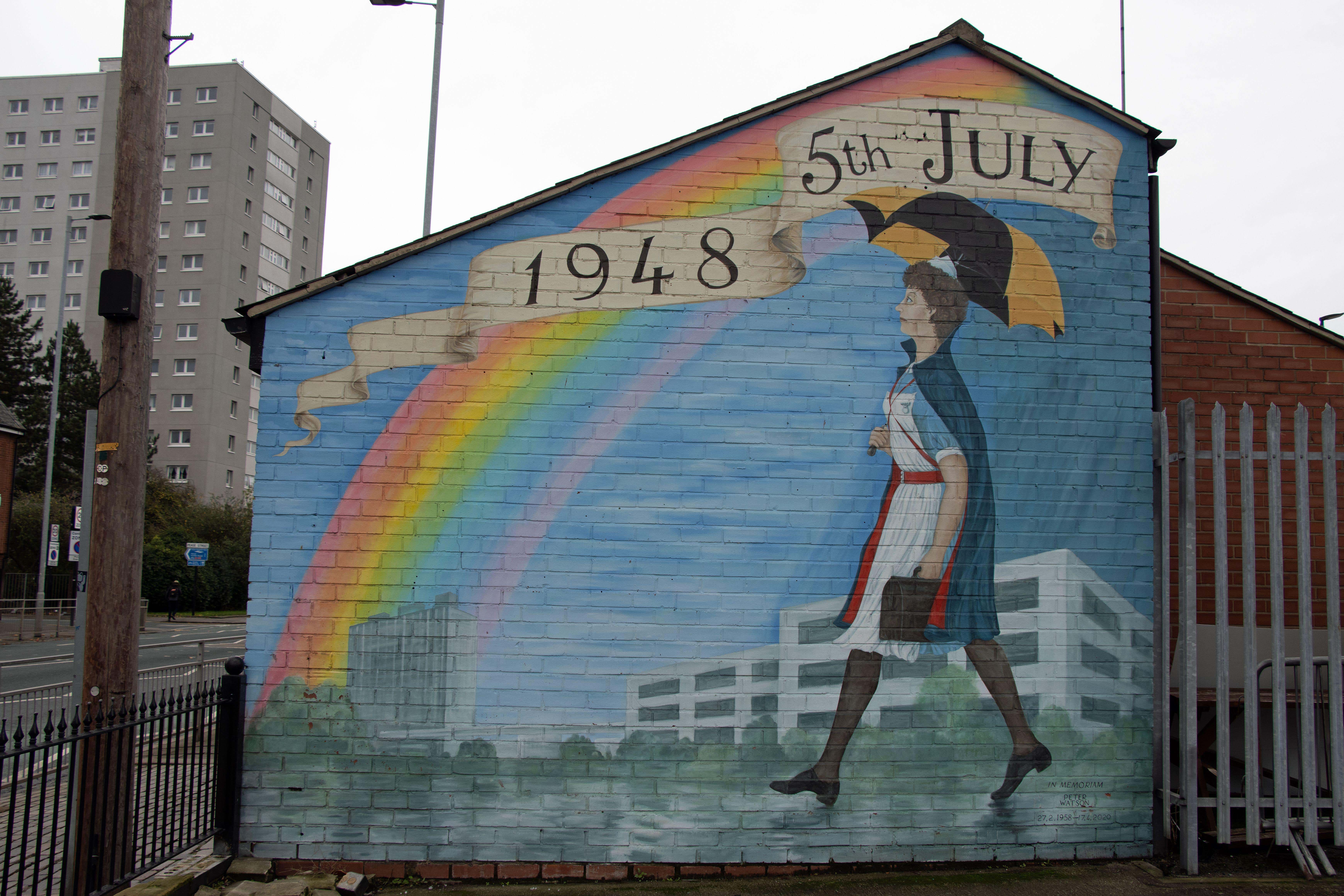 A mural on the side of a building.
The painting: There is a blue sky with a rainbow, there are buildings in the background, in the foreground a nurse walks across in an old fashioned uniform, holding an umbrella.