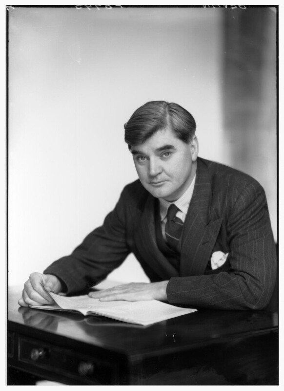 A black and white photograph of a white man sat at a desk with a book. He is staring at the camera. He is wearing a three piece suit with a tie. He has short, neat hair.
