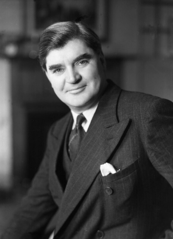 Black and white photograph of a white man. He is wearing a three piece suit and tie. He has short dark hair. He has a slight smile.