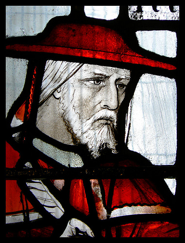 Stained glass. The shoulders and head of a white man (John Morton) who has white hair and a white beard and moustache. He is wearing a red hat and red robes. He looks solemn.