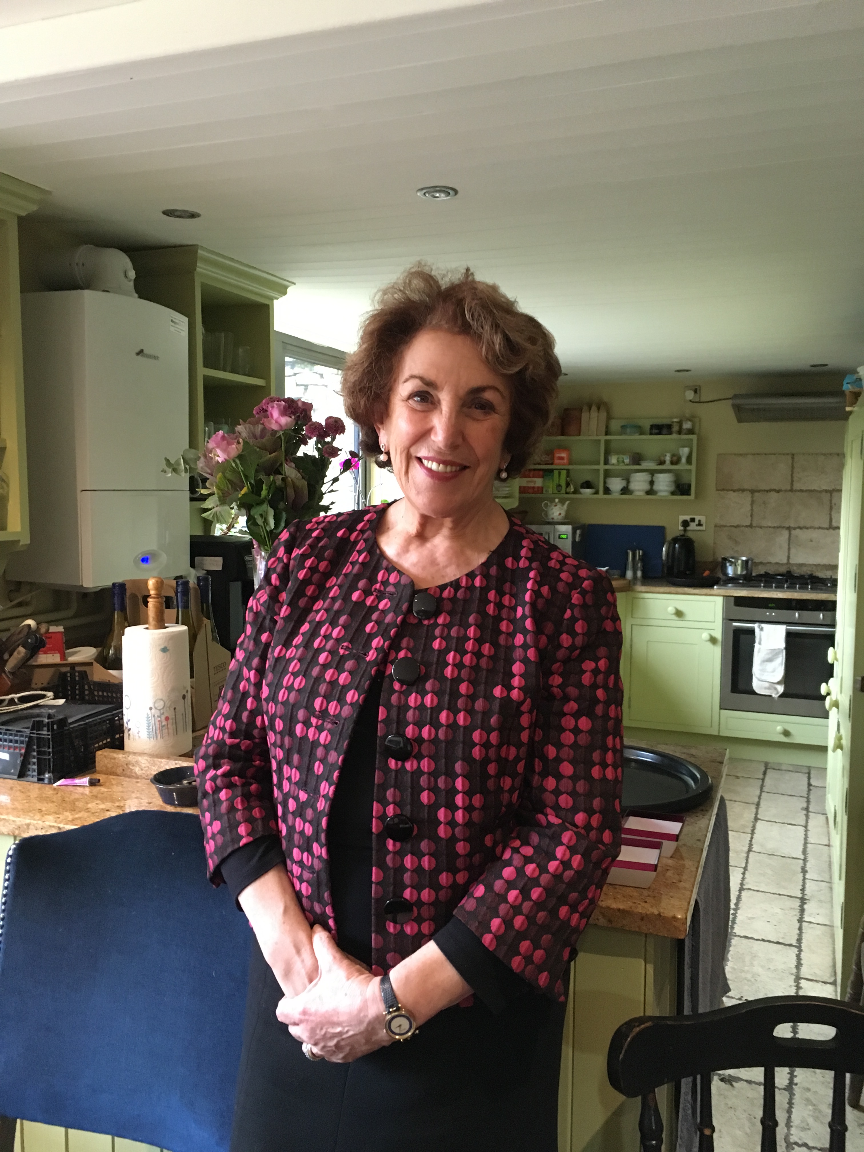 A photograph of a white woman stood in a kitchen. She has her hands clasped in front of her. She is wearing a black dress, a patterned jacket and a watch. She is smiling.