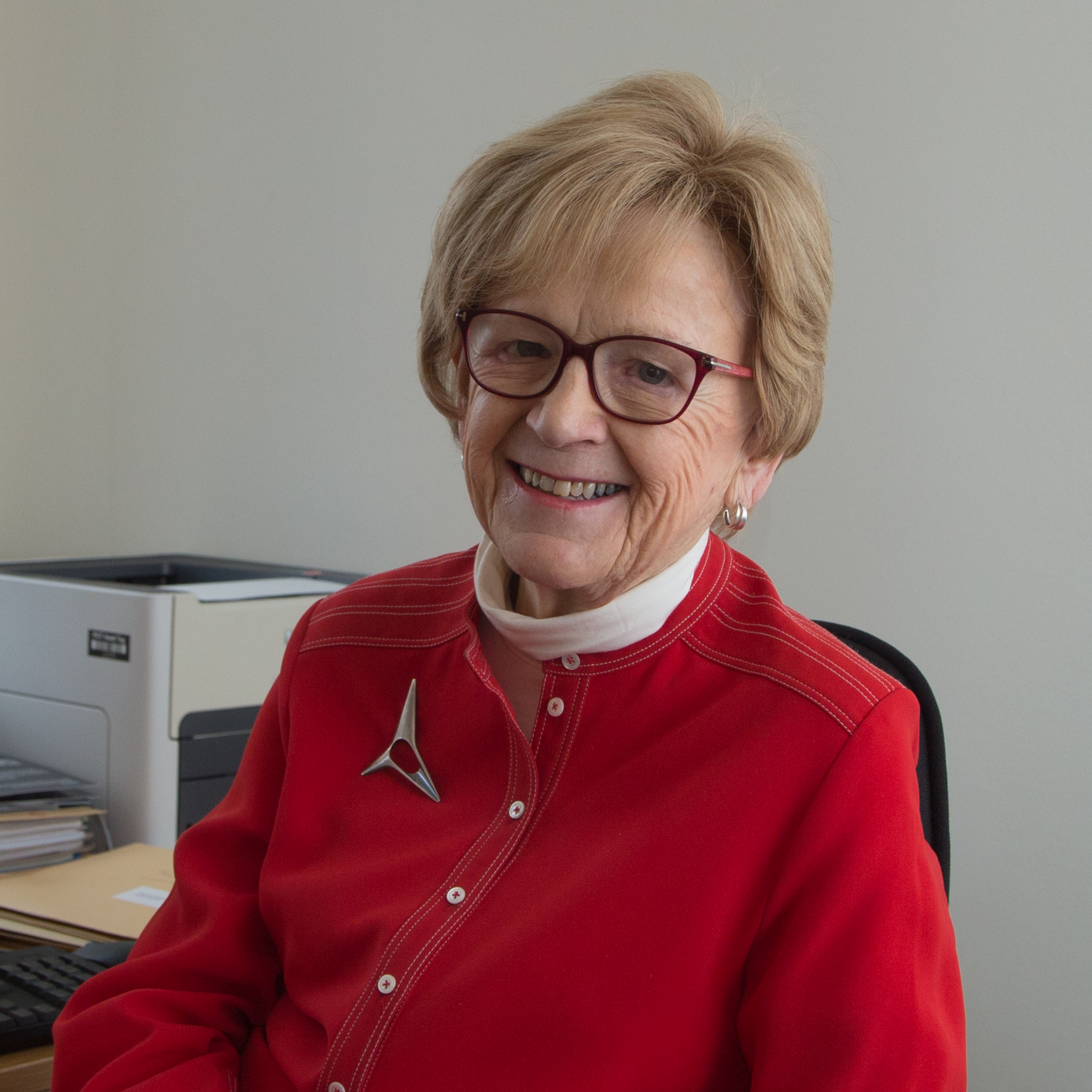 A photograph of a woman with short blonde hair. She is wearing a white top, red jacket, a brooch and glasses. She is smiling.