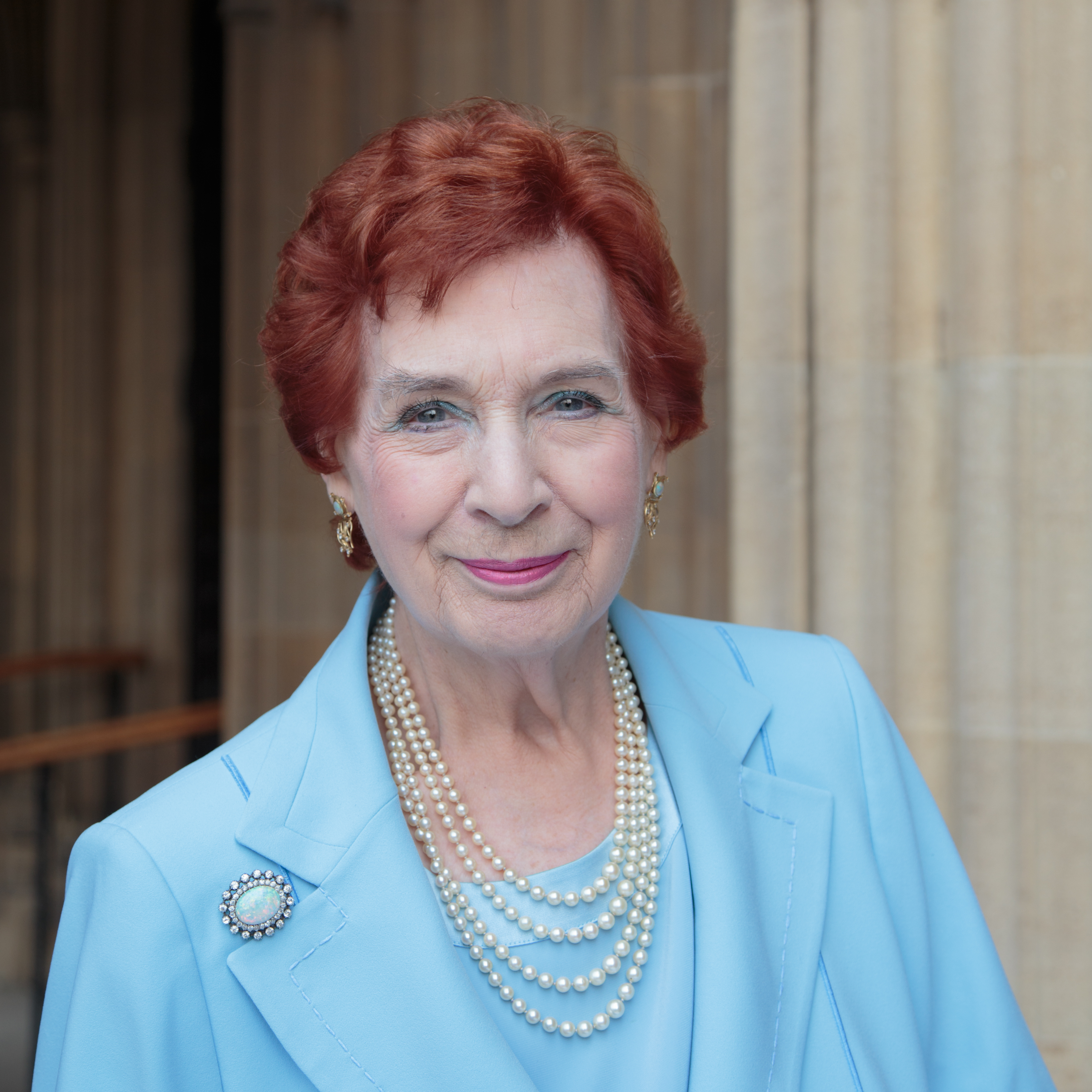 A photograph of a white woman with short red hair. She is wearing a blue top and a bluejacket, pearls and a brooch, and blue eyeshadow. 