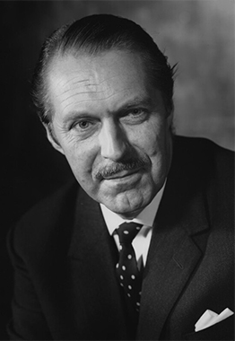 A black and white photograph of a white man with short thinning hair. He is wearing a shirt, jacket and spotted tie. He has a moustache.