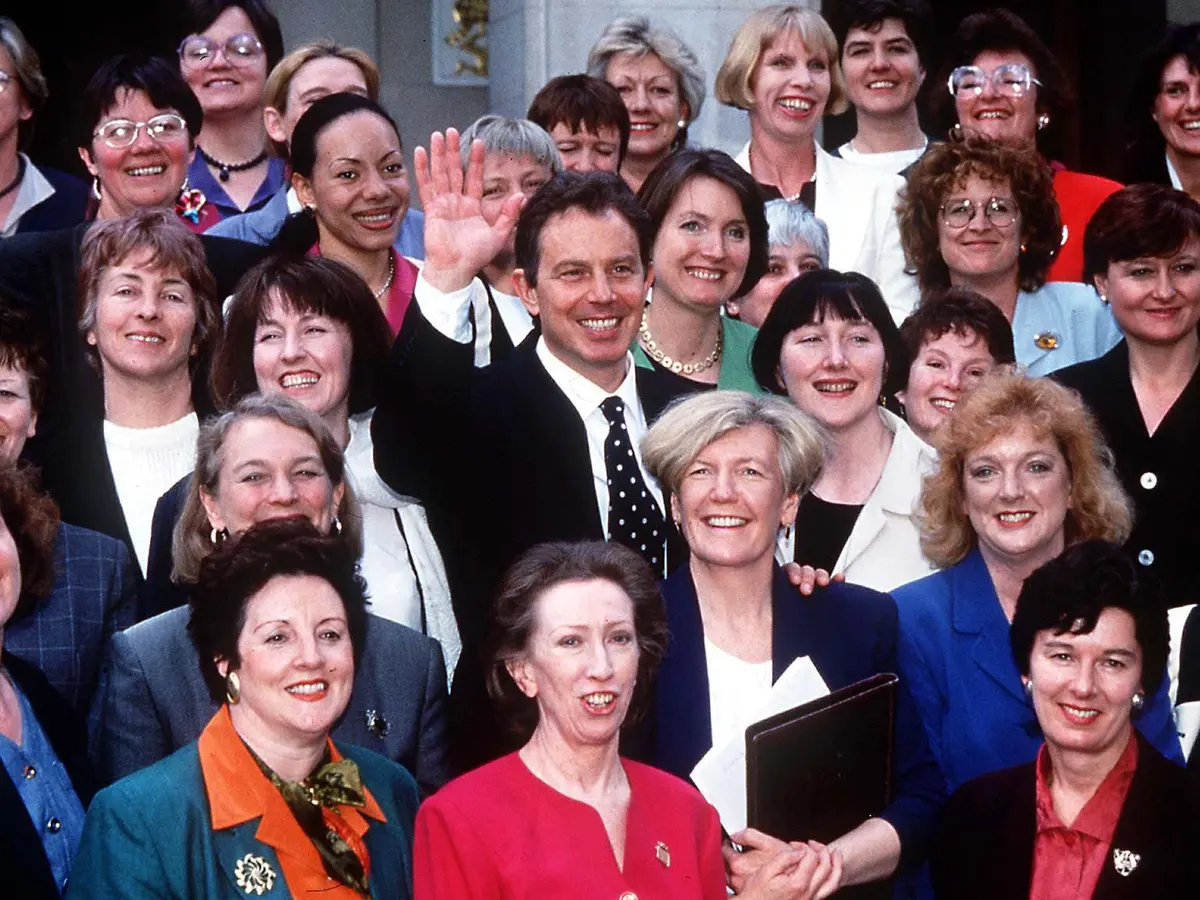 A photograph of a group of women smiling at the camera. In the middle of this group of women is a man waving. 
