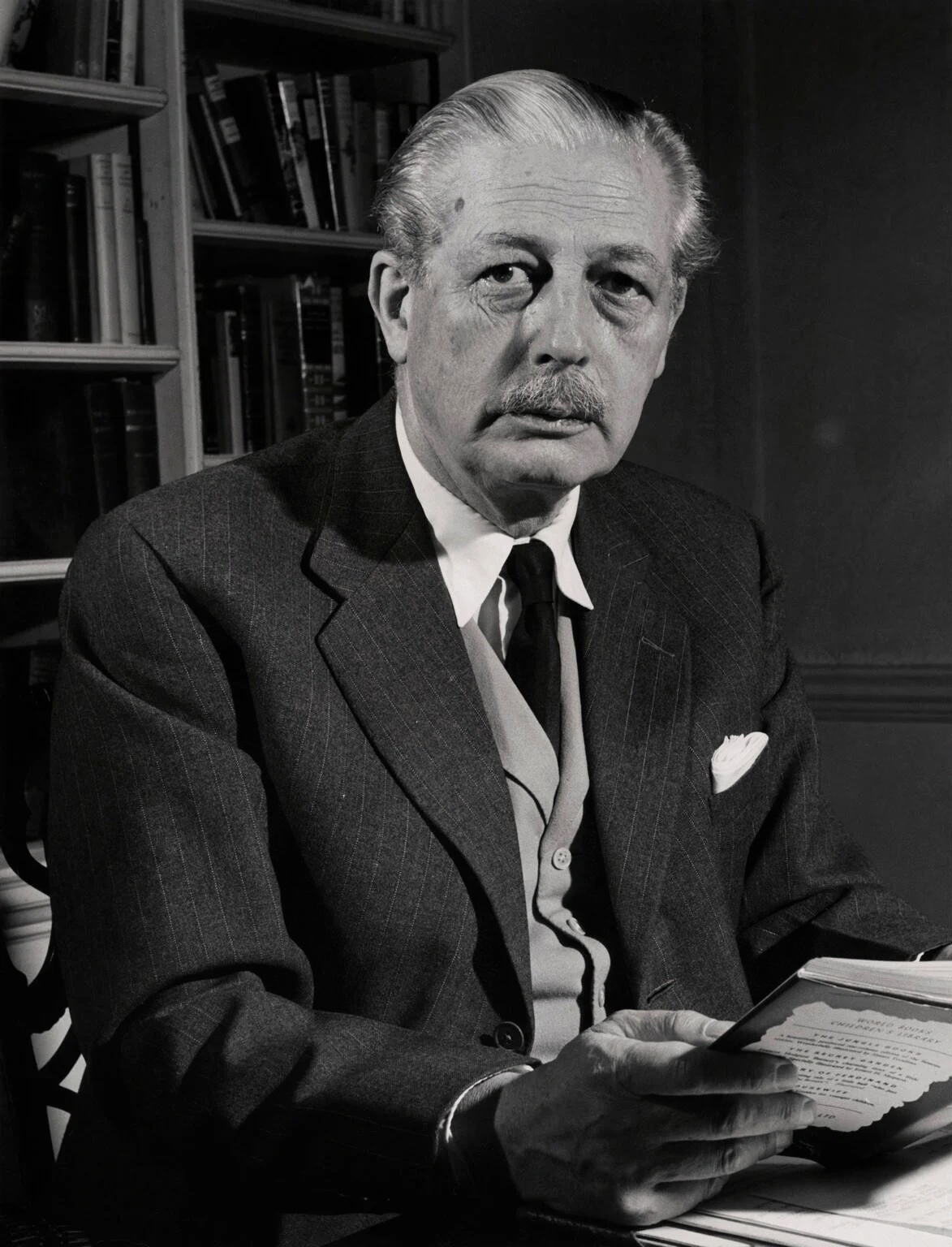 A black and white photograph of a white man with short hair and a moustache. He is wearing a three piece suit and tie. He is sat down at a desk, there is a book case behind him and he is holding an open book. 
