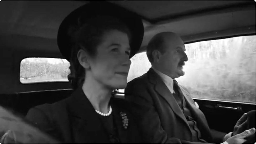 A black and white screenshot from a documentary. There is a man and a woman in a car.