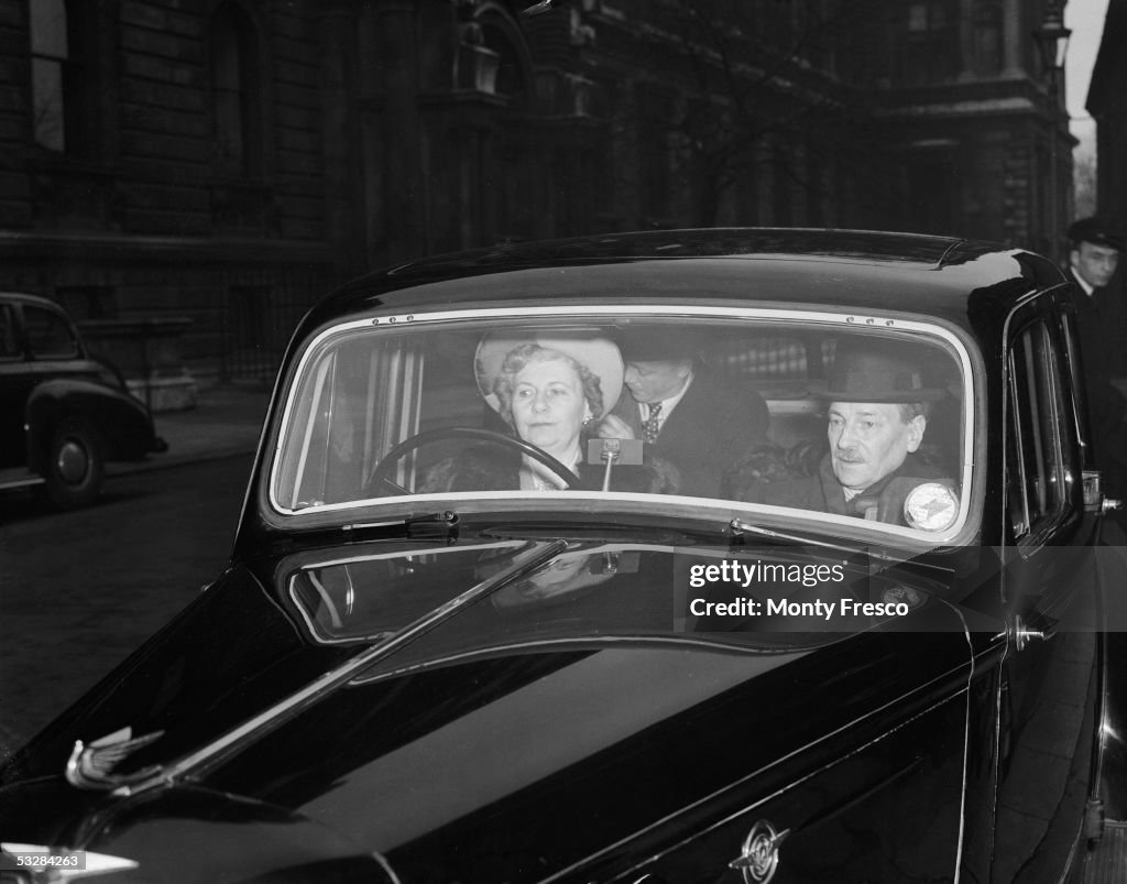A black and white photograph of three white people sat inside a dark coloured car. There is a woman driving the car wearing a hat, the other two passengers are men.