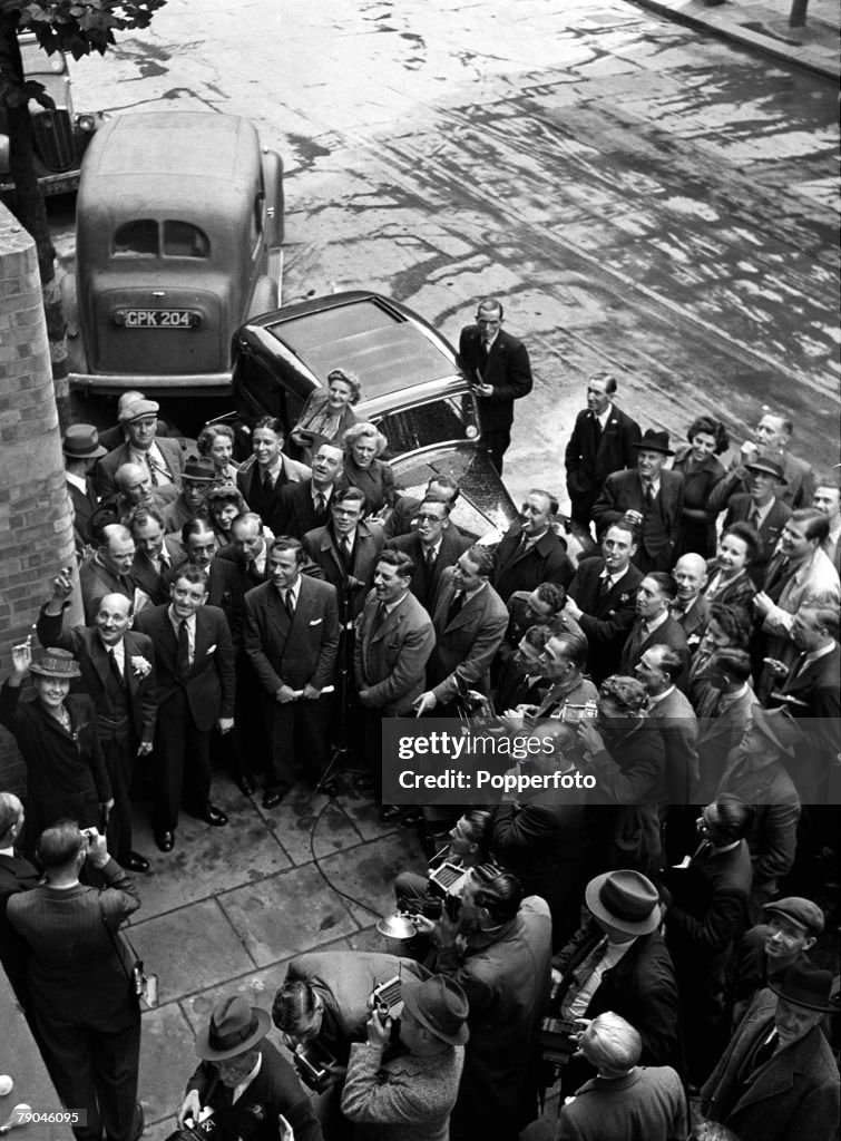 A black and white photograph of a crowd of journalists, photographers and people including two people (Mr and Mrs Attlee) waving towards the sky. There are cars parked in the background.