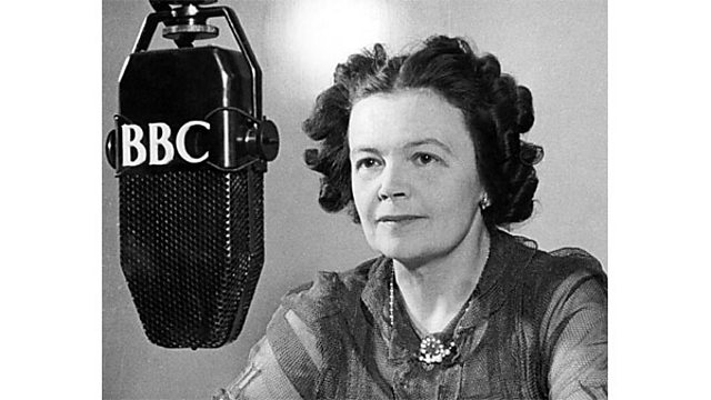 A black and white photograph of a white woman with short dark curled hair. She has a necklace and earrings on. She is sat beside an old school radio microphone that has the letters BBC on it.