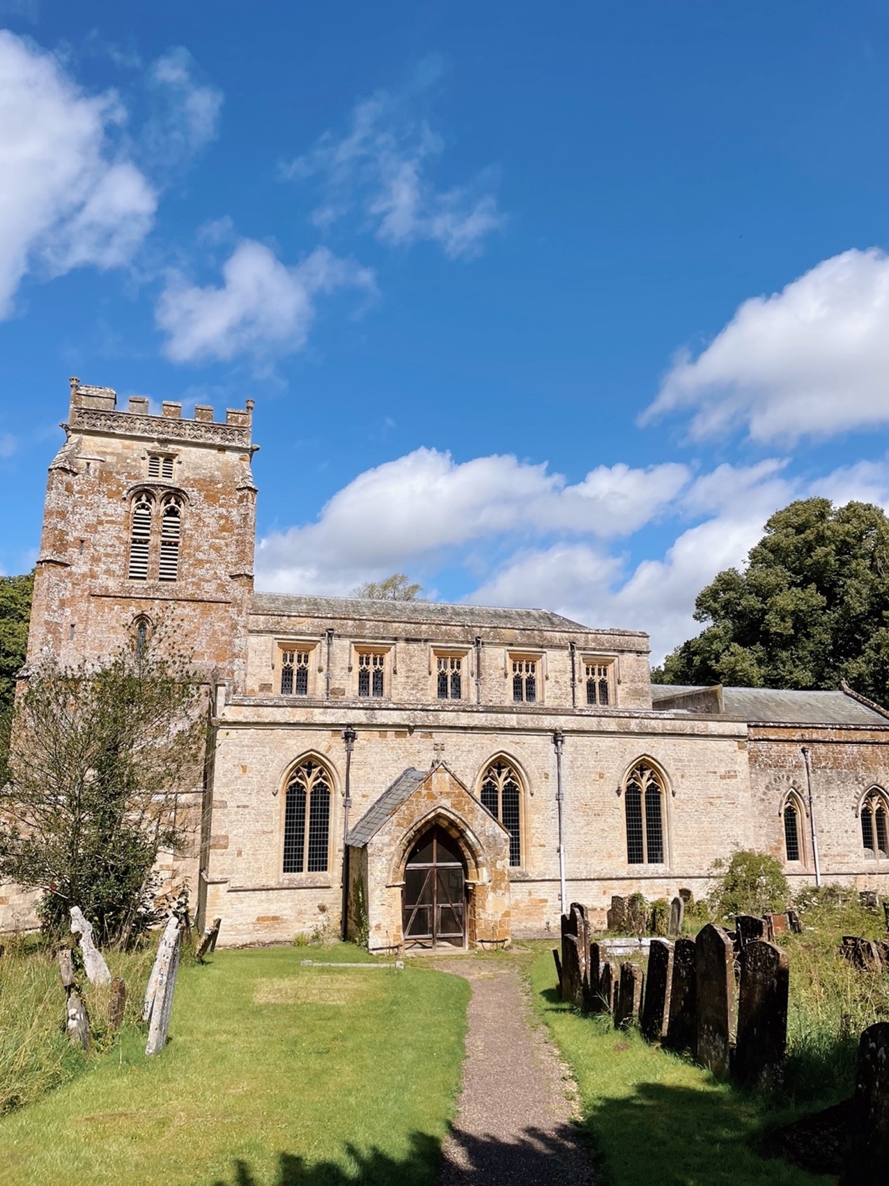A coloured photograph of a small church sat in a cemetery. The sky is blue with minimal white clouds ad there is a tree behind the church and one in front. There is a path in the middle of the cemetery leading to the doors of the church.