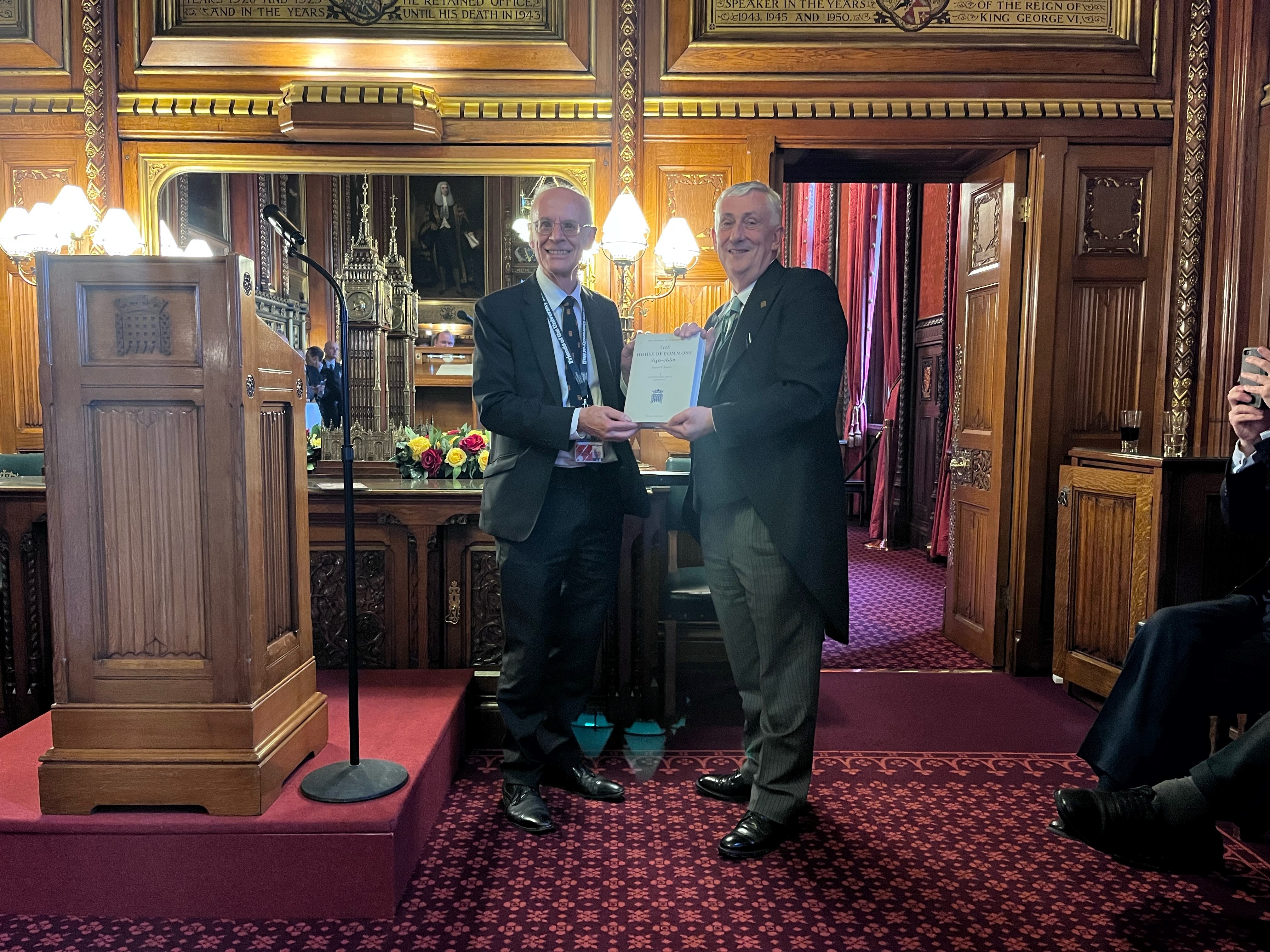 Two men, one in a black suit and the other in a mourning suit, both holding a book. The book has a cream cover. On the left of the image is a wooden lectern and microphone, the floor is covered with red patterned carpet and the walls are covered in wood panelling.