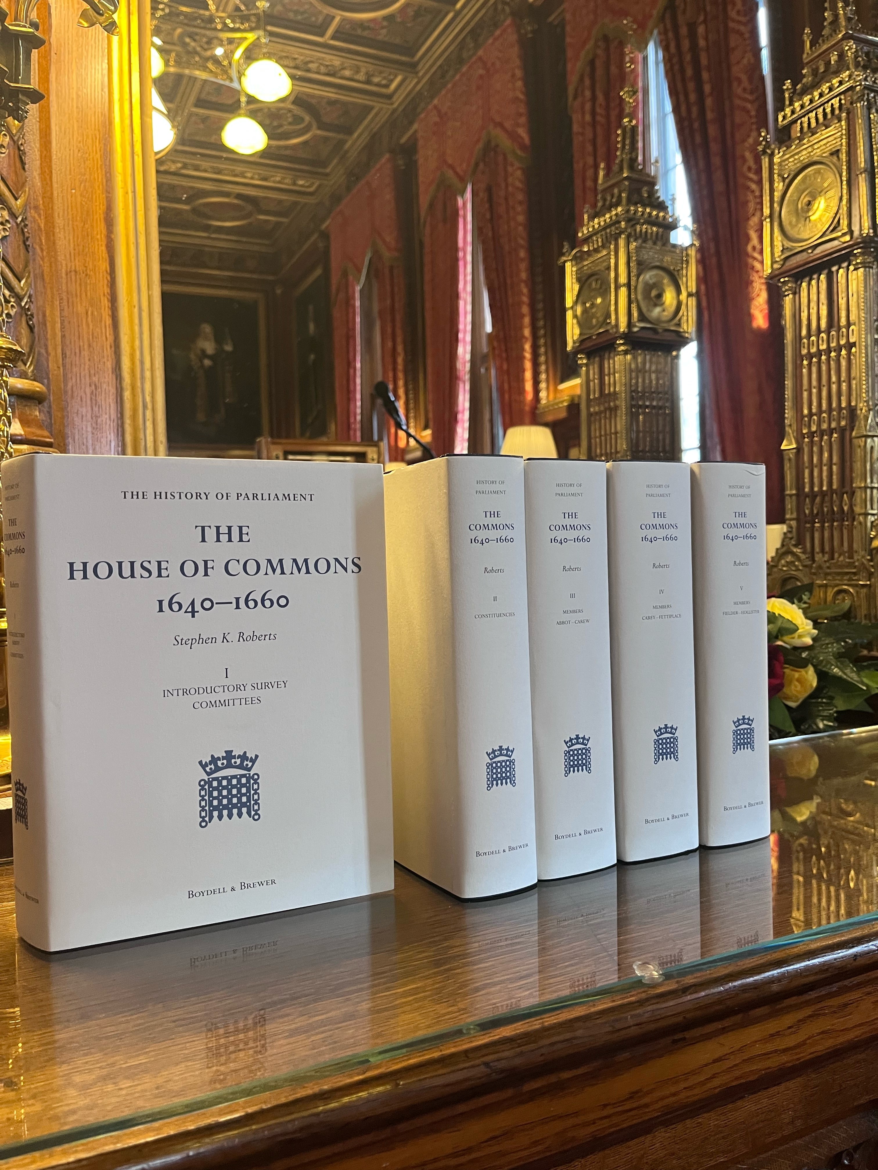 Five books with cream covers are stacked next to each other on a wooden surface. One book is arranged facing the camera, the cover reads 'The House of Commons 1640-1660'. A gold statue of the Big Ben clock tower is on the right had side of the image.
