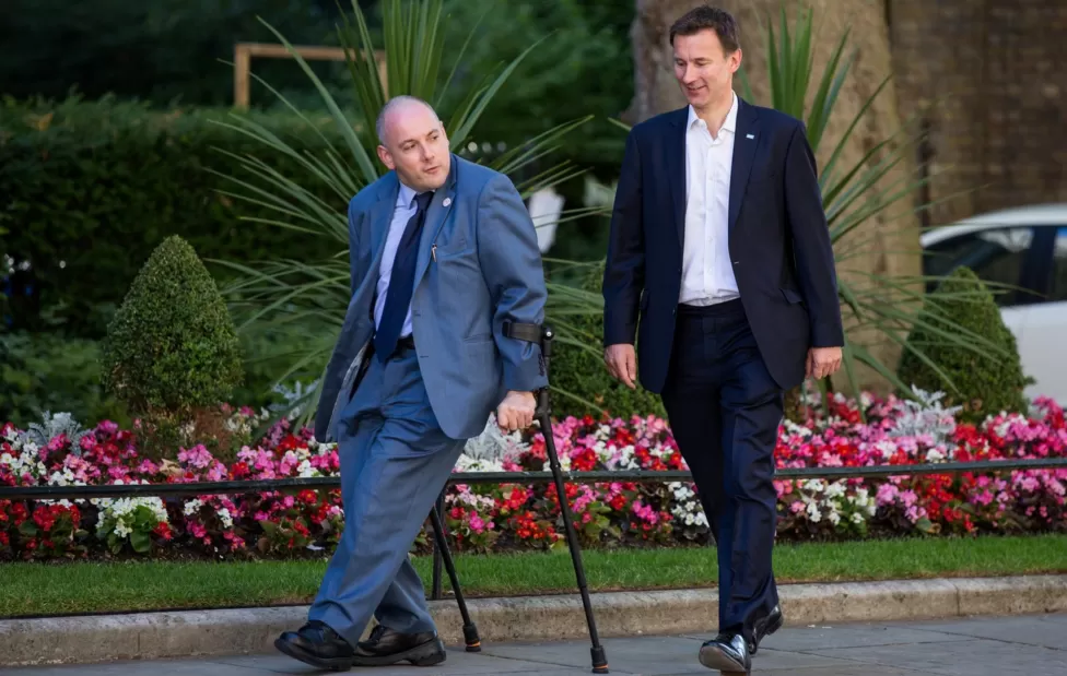 A photograph of two white men walking together. There are pink, red, and white flowers. The man on the left is wearing a blue suit and blue tie, he is walking with crutches, the man on the right is wearing a dark suit with no tie. They look like they're talking to each other. 