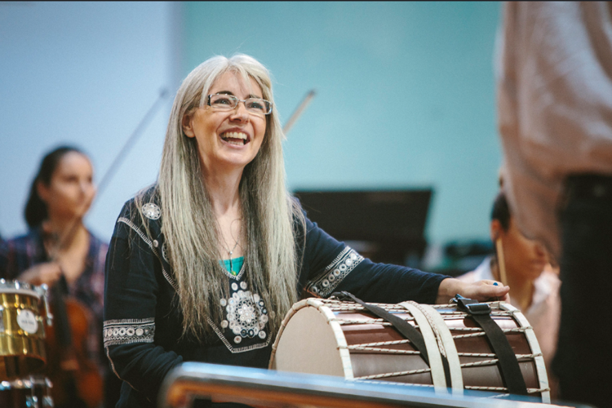 A photograph of a white woman with medium to long length white hair with bangs. She is wearing a blue shirt decorated with sequins. She is holding a large drum and smiling/laughing with an instructor out of shot. Around her are a few more people with different instruments, including a woman with a violin.