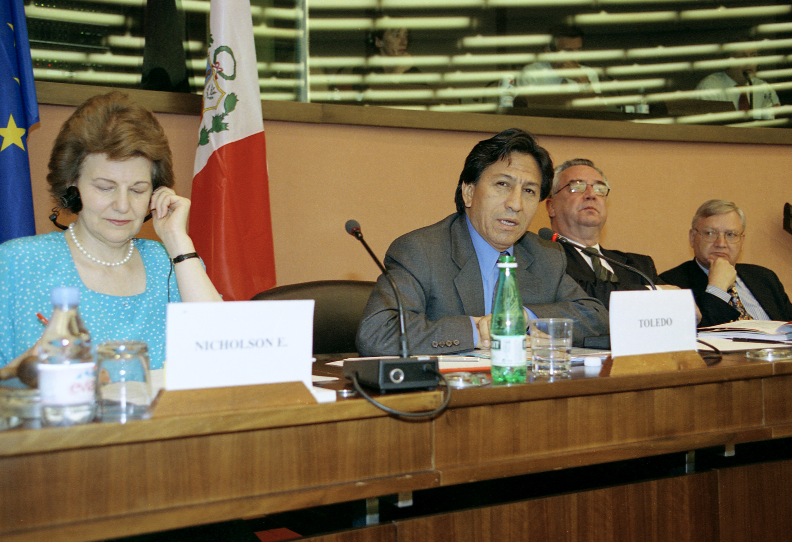 A photograph of four people sat behind a desk. There is one woman and three men. The men are all wearing suits and ties and two of them are wearing glasses. The woman is wearing a pearl necklace, a blue dress patterned with white and is holding headphones to her ears. On the desk are name signs, microphones and bottles of water with glasses. Behind the people is the European Union flag (blue with yellow stars in a circle) and the Peruvian flag (A vertical triband of red with white in the middle that has the National Coat of Arms centred on the white band.)