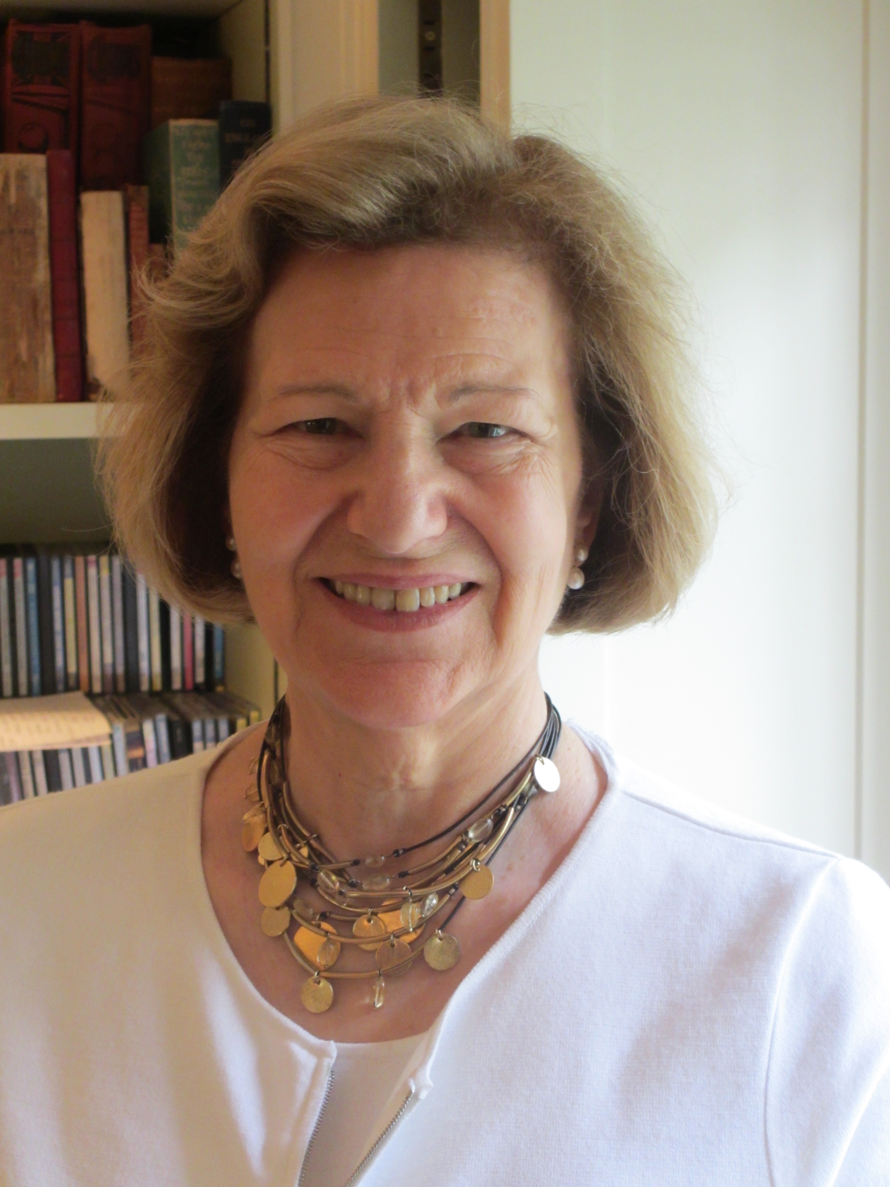 A photograph of a white woman stood in front of a bookshelf. She has chin length blonde/brown hair and is wearing earrings and a necklace. She is smiling at the camera.