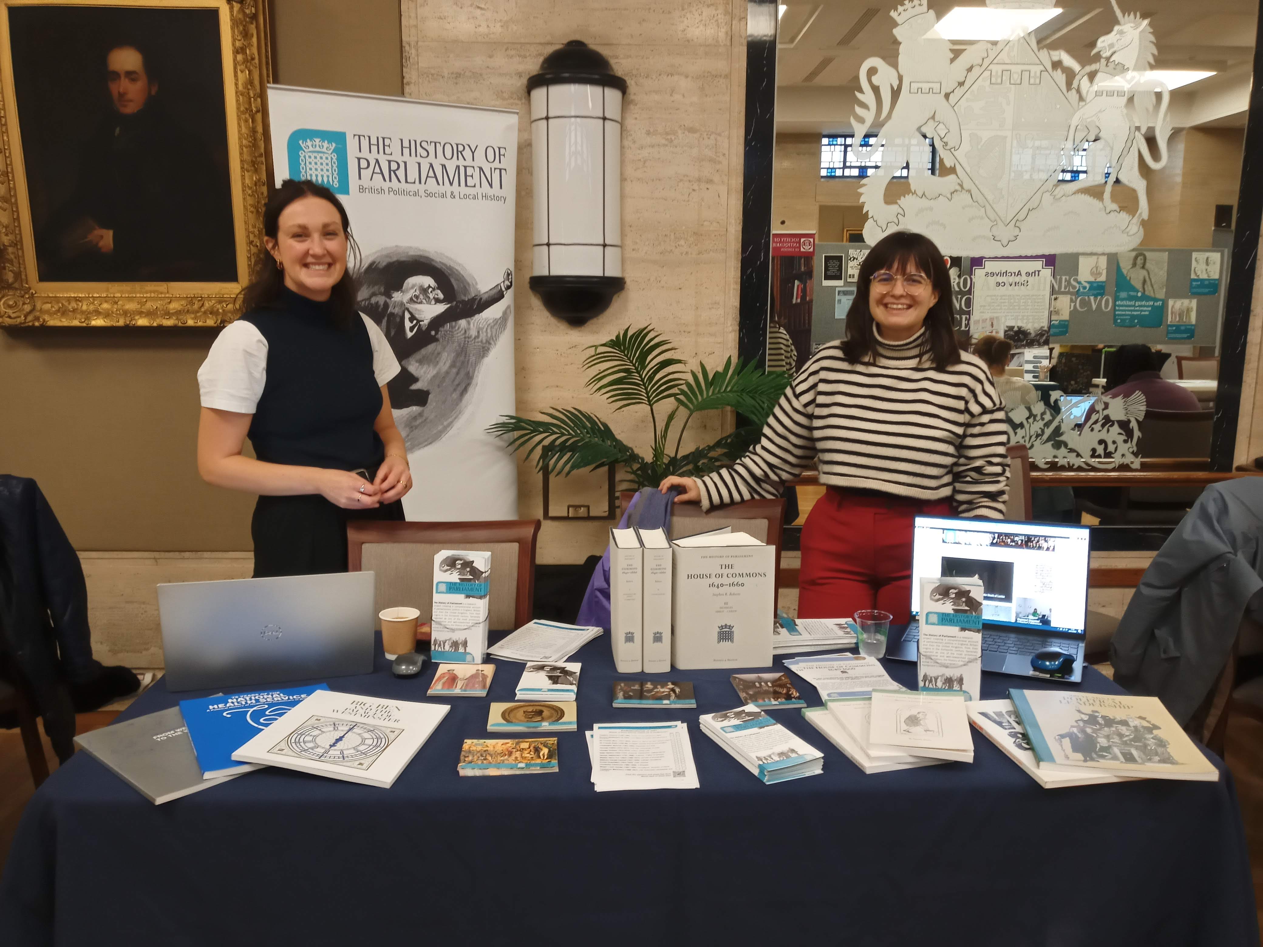 Two white women stand smiling at the camera. In front of them is a table, covered in a navy tablecloth, displaying a number of books and leaflets. A banner behind them reads 'the History of Parliament'.