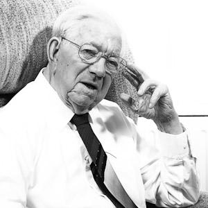 A black and white photograph of a white man with thinning white hair. He is wearing glasses and a shirt and tie. He is sat down on an armchair and is leaning on his left hand. He is looking at the camera with a serious expression.